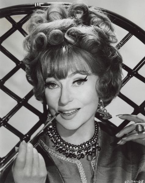 Quarter-length portrait of Agnes Moorehead as 'Endora' from the TV show "Bewitched." She is holding a cigarette holder up to her mouth and is smiling. The chair she is seated in has a high, latticed back. The photograph is part of a publicity packet from April 5, 1968.