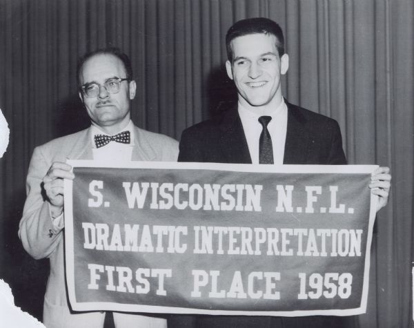 A teenage Daniel J. Travanti stands next to a man, with the two men holding a banner that reads: "S. Wisconsin N.F.L., Dramatic Interpretation, First Place 1958." N.F.L. stands for National Forensic League.