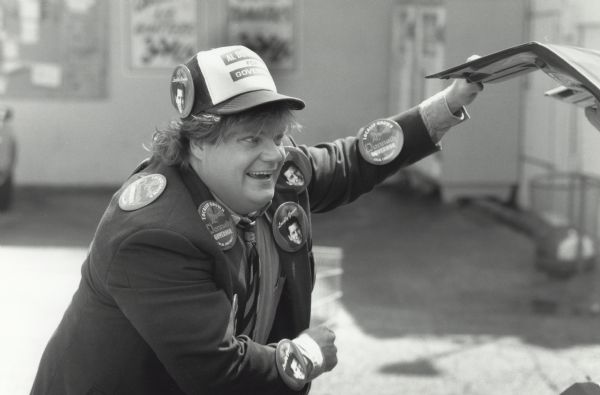 Chris Farley's character in the movie <i>Black Sheep</i> looks into a car trunk. He is wearing a suit with a baseball cap, and his suit and cap are covered with many large political buttons.