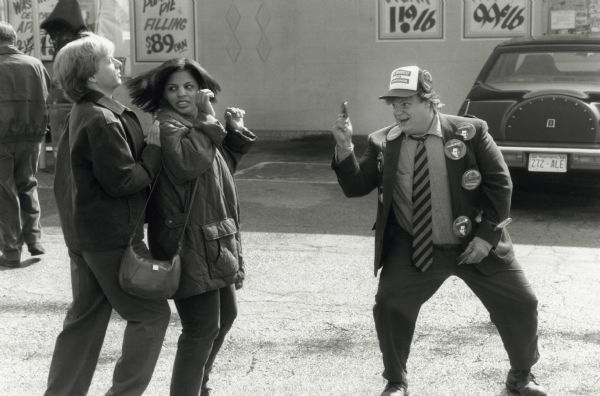 Chris Farley's character in the movie <i>Black Sheep</i> enthusiastically offers a political button to a man and a woman. They are standing in a parking lot. Chris Farley is wearing a suit with a baseball cap which is covered with a number of political buttons.