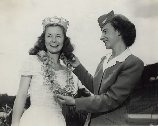 Margaret McGuire receives a lei made of orchids in a ceremony making her the Queen of the Wisconsin Centennial Exposition at the State Fair. The lei was flown in from from Hawaii for Governor Ingram Stainback of Hawaii and presented by Mary Ludwig, airline hostess, in uniform. Miss McGuire is wearing her Alice in Dairyland crown and served as hostess of the Dairy Building. 