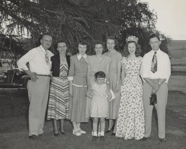 Margaret McGuire (wearing the crown) and her family, with the photographers, Mr. and Mrs. Edgar Obma, posing outdoors on the lawn at the Dodgeville-Mineral Point Country Club. A note on the back reads, "Michael (nap time!)," probably referring to the little girl. In the background are trees, fields and a parking lot with automobiles.
