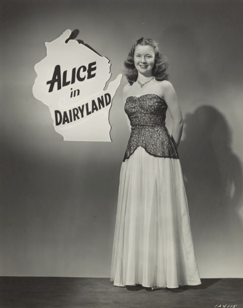 Margaret McGuire, winner of the Alice in Dairyland contest at the Schraeder Hotel. She is wearing a light-colored, floor length, strapless dress with dark lace over the bodice, waist and hips. She is posing next to a sign in the shape of the state of Wisconsin. Text on the sign reads: "Alice In Dairyland."