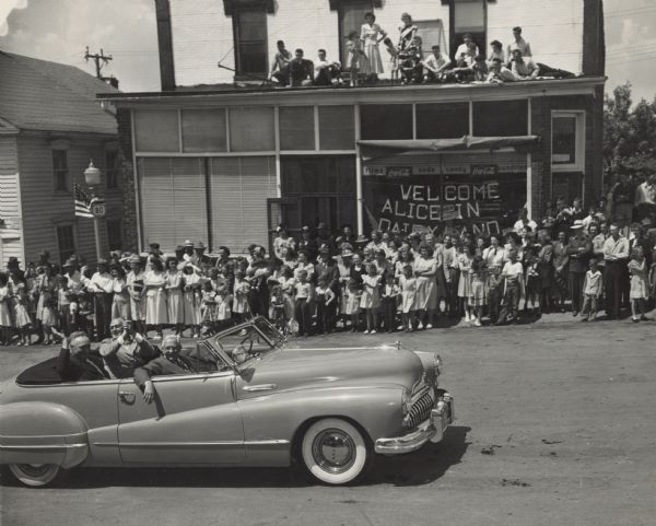 Elevated view of Governor Rennebohm, sitting in the front passenger seat of a convertible. He is riding in the parade to honor the first Alice in Dairyland. Spectators line the opposite side of the street, as well as from the roof of a storefront. A banner in the show window reads: "Welcome Alice in Dairyland."