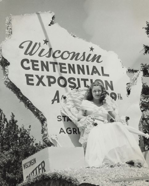 Margaret McGuire, Alice in Dairyland, sitting in chair decorated with fabric and flower on a float in a parade. She is wearing a light-colored, full-length dress, and is holding a parasol in her lap. Beside her is a box labeled: "Wisconsin Butter." Behind her is a cutout of the state of the shape of Wisconsin with the text: "Wisconsin Centennial Exposition, August 7th-29th, State Dept. of Agriculture."