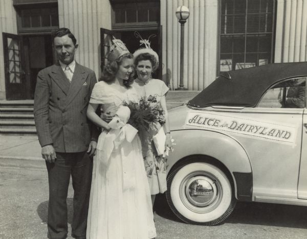 Margaret McGuire, Alice in Dairyland, standing between a man and woman next to an automobile. The woman has her arm around Margaret's waist. Margaret is wearing a full-length dress, a crown on her head, and is carrying a bouquet of flowers. The automobile, a Chevrolet convertible with the top up, has a banner that reads: "Alice in Dairyland" above the rear fender.