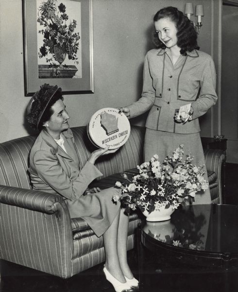 Margaret McGuire, Alice in Dairyland, posing standing, with a woman sitting on a sofa at the Barclay Hotel. Between them they are displaying a round gift tin of cheese. The text on the tin reads: "From America's Dairyland, Assortment of Fine Wisconsin Cheese, Packed by Gimbels Milwaukee." The room has a sofa, round table with flower arrangement and a painting of a vase of flowers on the wall. Both women are wearing suits with skirts.