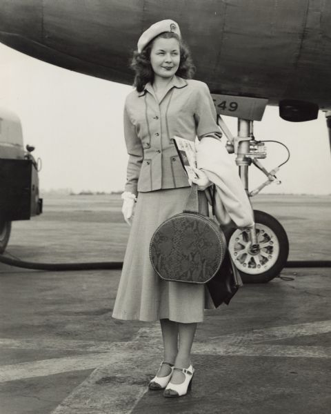 Margaret McGuire, Alice in Dairyland, standing on the runway immediately after her arrival. She is wearing a suit with a skirt, hat and gloves. In her arm is a folder, coat and carry on bag. In the background is the nose of the airplane, the runway and a the back of a fuel truck.