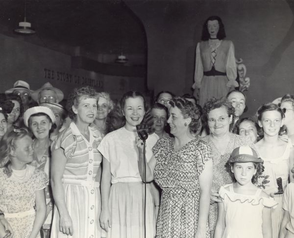 Alice in Dairyland, Margaret McGuire, speaks into a WHA microphone in the Dairy Building at the Wisconsin Centennial State Fair. She is surrounded by a crowd of mostly women and girls. In the background on the left the words: "The Story of Dairyland" are mounted on the wall. On the right is the "Mechanical Alice." After Margaret won the competition, the Dairy Committee decided to have a 10-foot tall mechanical version made of her as Alice. Sculptor John Neidhart created the statue's head and based its features on Margaret. The rest of the body was built in Pittsburgh, Pennsylvania. Upon its completion, the mechanical Margaret was dressed in the same "Alice in Dairyland" prize-winning costume. Besides raising her arms and turning her head, the statue could also sit down on her oversized throne.
