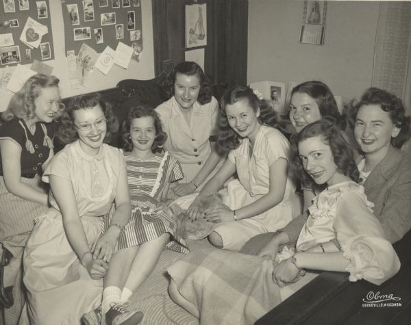 Margaret McGuire, Alice in Dairyland, (5th from left), with six of her roommates and the House Mother, Ms. Welch, at Whitewater State University. They are seated on her bed, all wearing dresses. Margaret holds a stuffed animal. Above the headboard are two bulletin boards with photographs and keepsakes pinned to it.