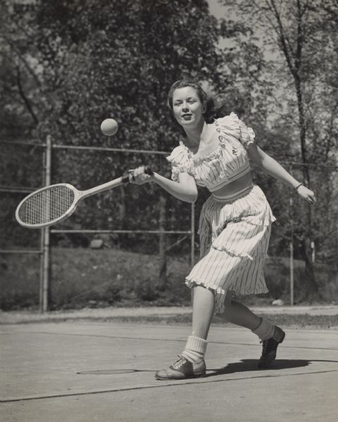 Margaret McGuire, Alice in Dairyland, playing tennis outdoors at Whitewater State University in her Physical Education class. She is wearing a skirt, midriff top, saddle-shoes and bobby socks. In the background is a fence, lawn and trees.