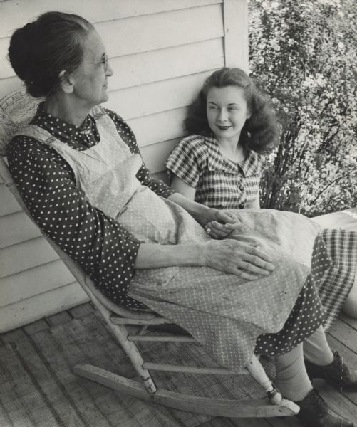 Margaret McGuire, Alice in Dairyland, sitting on the porch floor behind her Grandmother, Mrs. Christine McGuire, who is sitting in a rocking chair. In the background on the right is a flowering shrub. They are both wearing dresses and her Grandmother is wearing an apron.