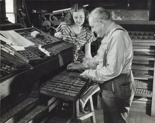 Margaret McGuire, Alice in Dairyland, is perched on a stool watching a typesetter at <i>The Highland Press</i>. He is setting metal type by hand for the weekly newspaper. They are surrounded by type cases, type cabinets and printing machinery. He is setting type into a composing stick in his hand and the composed type is in a gallery in front of him.