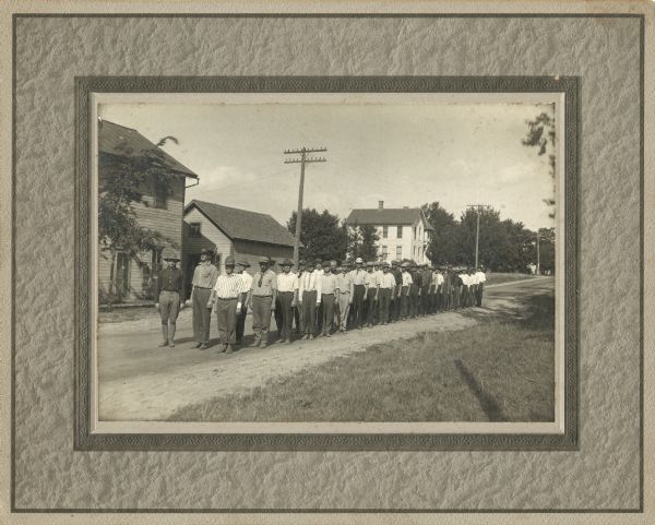 Men from Company "C" are lined up in a double row on a dirt town street. A soldier in uniform stands at the head of the line. In front are Ed Fryk (2nd from left) and Herbert Weinmann (3rd from left). It is possible that they have just enlisted. In the background are homes, trees and power poles.

Question: Name of town?