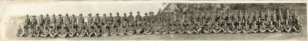 Panoramic group portrait of Company "K" Fourth Wisconsin Infantry. In the background is a rock formation, one of several in the area, with trees. The soldier (12th from the left in the back row) is Herbert Weinmann. Text at the foot of the photo identifies "Capt. C.J. Rollis, 1st Lieut. E. Wood and 2nd Lieut. C.A. Rowe" but does not indicate who they are. They are wearing uniforms and holding their weapons.