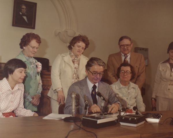 Kathryn Morrison (far left) observes as Governor Patrick Lucey signs, possibly, a piece of legislation. State Representative Mary Lou Munts (seated next to Lucey) is also looking on.