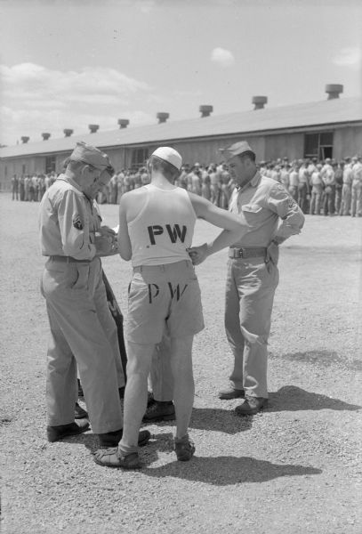 Two German prisoners and two American officers discussing some papers while standing outdoors. In the background, German prisoners of war are lined up for inspection outside their barracks before going to work at a local cannery.