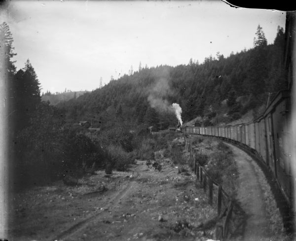 View alongside railroad cars towards the locomotive in the distance, which is traveling along a curve in the tracks. There are a few buildings near the railroad tracks at the base of forested hills.  At this time Carl Peterson was writing letters home to Christine Jenson about his journey.