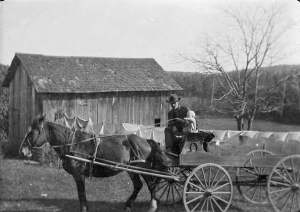 View across yard towards Carl Peterson sitting next to his son James holding the reins of two horses while sitting on a wagon. In the background are farm buildings and laundry hanging on a clothesline.