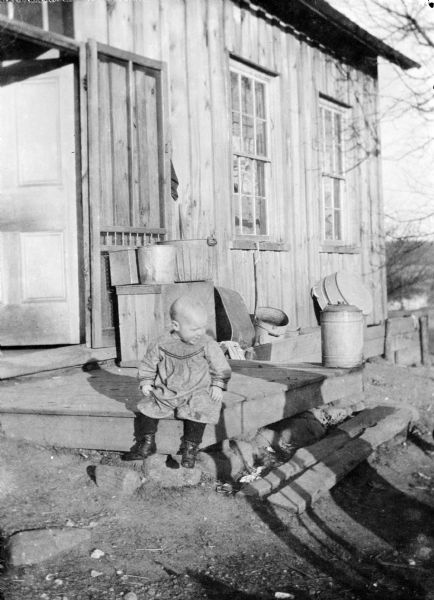 View across the yard towards James Peterson sitting on a landing near the open door of a house or farm building with two windows. Stacked on a crate near the door are buckets, and other containers and a washtub are along the side of the building.