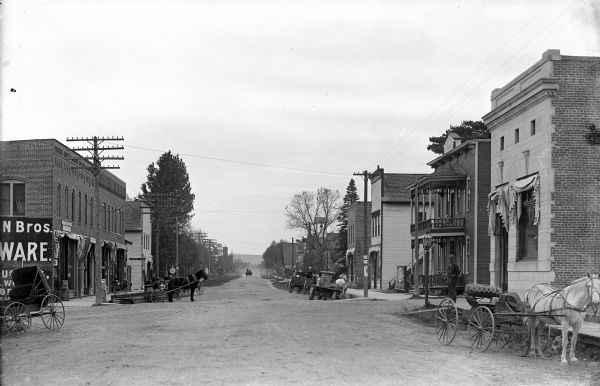 View down center of an unpaved Main Street with commercial buildings along sidewalks. Horse-drawn vehicles are parked near the curb and moving further down the street in the far distance.