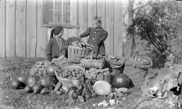 Laurie and Muriel Peterson are standing behind a display of farm produce from their mother's garden. On the ground in front of them are pumpkins, squash, and apples and potatoes and other vegetables in baskets and sacks. Behind the children is a wood sided building with a window.