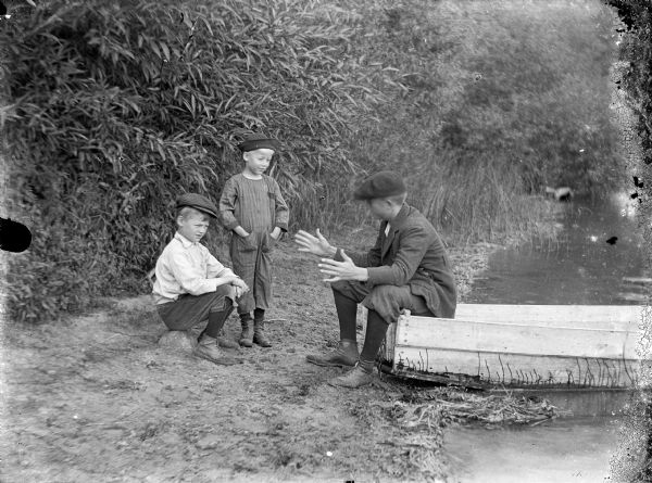 View towards Laurie Peterson sitting on a boat pulled up on the shore, gesturing with his hands spread apart, telling a story to his two cousins, Alton and Arthur, listening on the bank. In 1919, Carl and Christine took in Alton and Arthur after their mother passed away in the influenza epidemic.