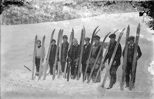 Scandinavia Ski Team, all men, are posing in a row with their skis propped on their shoulders. In the background is a snow-covered hill and trees.