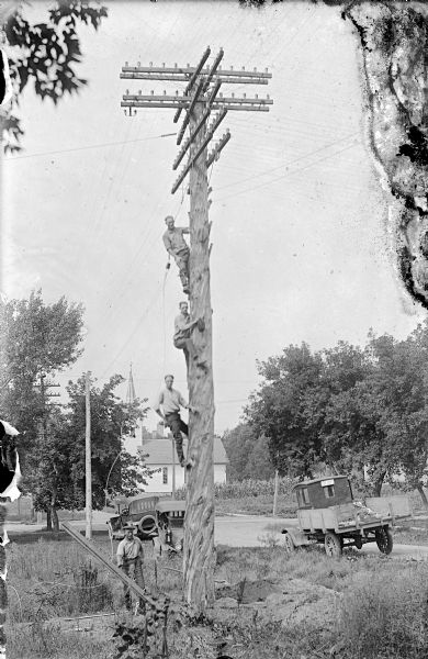Group portrait of four telephone linemen. Three of the men are posed on the pole, and another man is standing on the ground. Three automobiles are parked along the roadside behind the men. In the far background is a church building. The man on the top is Carl Peterson and the third man down is Laurie Peterson, son of Carl. Carl was a troubleshooter for the telephone company.