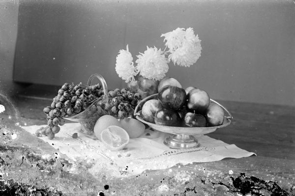 Still life with flowers in a vase, apples in a silver bowl, grapes in a glass basket, and oranges on a white cloth.