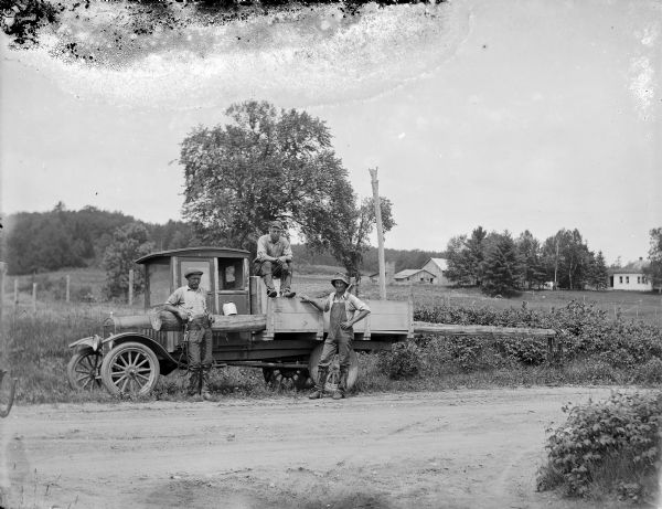 View across dirt road towards three linemen posing in front of a truck that is parked along the side of the road. A power pole is attached horizontally in a special holder attached to the truck box. In the background are fields and farm buildings.