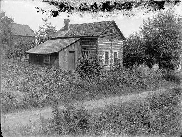 Log house with lean-to addition. There is another building in the background on the left. A road or path is in the foreground. Carl and Christine Peterson lived in this cabin from 1908 to 1916. Previously, they lived with Christine's mother.