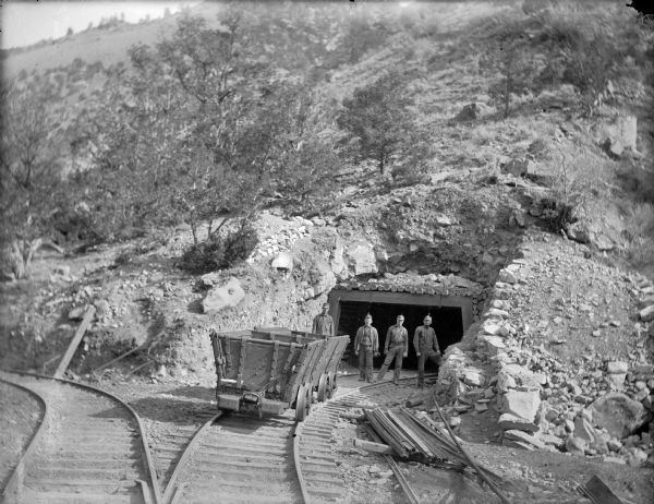 View down set of two railroad tracks towards four coal miners posing in front of a mine entrance near a coal car. The caption in the book reads, "Strike-breaking coal miners in Utah." At this time Carl Peterson was writing letters home to Christine Jenson about his journey.