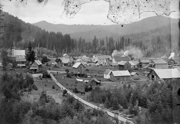 Elevated view from hill looking down on a mining town surrounded by wooded hills. There are wooden sidewalks between the houses. In the background on the left is a church building. At this time Carl Peterson was writing letters home to Christine Jenson about his journey.