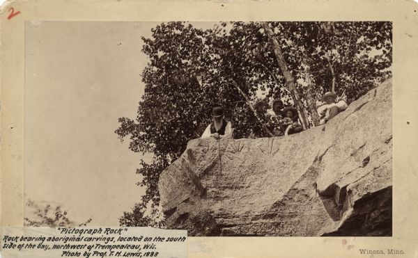 View looking up at three men and a child on top of a cliff on which are petroglyphs. Caption on photograph reads: "Pictograph Rock. Rock bearing aboriginal carvings, located on the south side of the bay, northwest of Trempealeau, Wis." Additional information on reverse, "Perrot State Park — these are petroglyphs destroyed by highway construction."