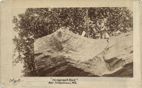 View looking up at three men and a child on top of a cliff on which are petroglyphs. Pictograph Rock bearing aboriginal carvings, located on the south side of the bay, northwest of Trempealeau, Wis." Additional information on reverse reads: "Pictographs, Trempealeau Rock Shelter, Plate 2. Perrot State Park — these are petroglyphs destroyed by highway construction."