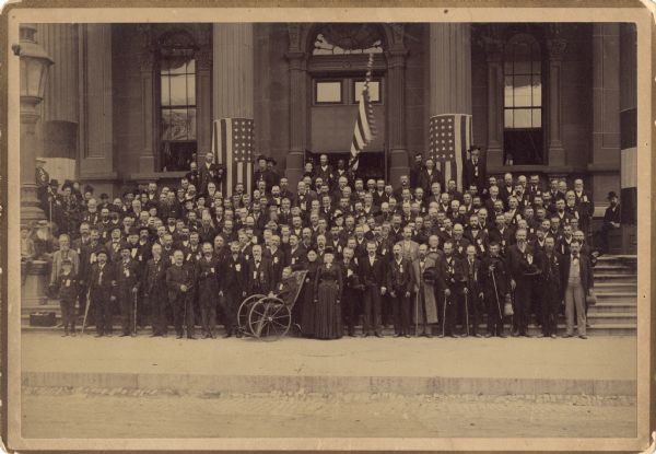 Formal outdoor group portrait of the 29th Wisconsin Infantry Volunteers. They are posing while lined up on the front steps of the Milwaukee Armory building. Two American flags are wrapped around the columns and one flag is being held by a soldier. Many soldiers have canes and one soldier is in a wheelchair.