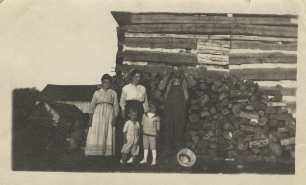 The extended Ott family posing in front of a woodpile of split and cut wood stacked against a log barn located on the Ott farm. The man is adjusting his glasses, and his hat is on the ground. Other farm buildings are in the background. Names are (back row, l to r): Anna Ott, Martha (Ott) Mensch (Martha's daughter and Gustave's step-daughter), 1893-1980, Gustave Ott, (front row, l to r) Martha's children: Rex Mensch (1917-1989) and Wilmer Mensch 1914-1958).