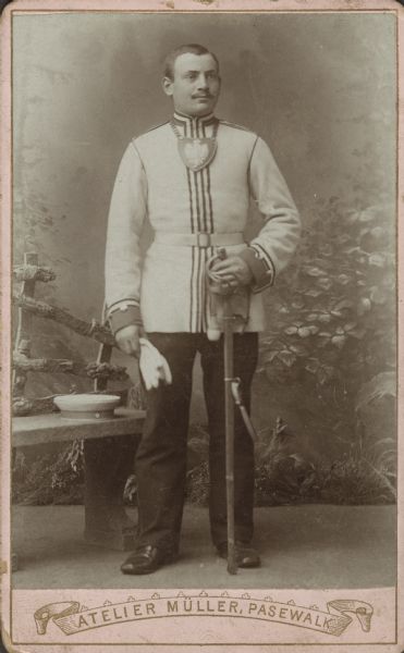 Formal studio carte-de-visite in front of a painted backdrop of Carl Wilhelm Berg. He is dressed in the uniform of the Prussian Heavy Cavalry, his rank is an NCO, probably a sergeant. He is holding his white gloves in one hand and his sword, in the scabbard, in the other. On the left his cap is resting on a stone bench. In the background is a prop fence and foliage. At the bottom of the card is the name of the photographic studio printed in a banner: "Atelier Muller, Pasewalk." Translated: "House of Muller, Pasewalk."<p>Carl Wilhelm Berg is Martha S. Bielke’s birth father. He remained in Germany and became a Major in the Kaiser’s Army.