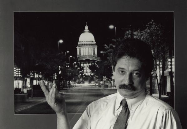 Portrait of Mayor Paul Soglin gesturing in front of a photograph of State Street looking up towards the Wisconsin State Capitol.