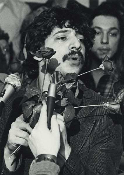 Paul Soglin, 27 years old, speaking into microphones while holding three roses. He is wearing a corduroy jacket. The location may be the Labor Temple on the evening of his election as Mayor over William Dyke.