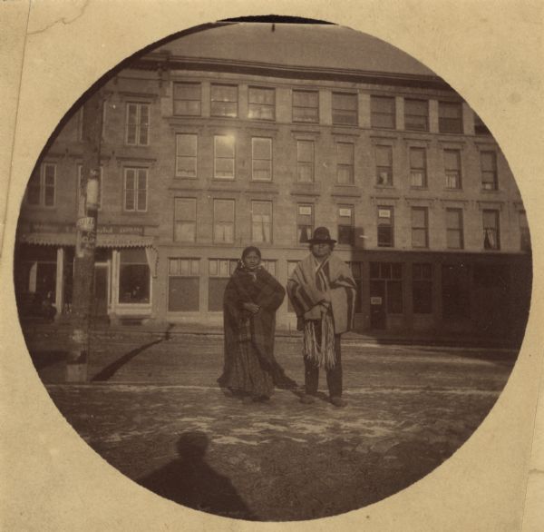 George Goodvillage and his wife posing on King Street. In the background is a row of commercial buildings. The building on the left is a Saddlery. The telegraph pole is on the left is covered with advertising posters, and a man's shadow is in the foreground.