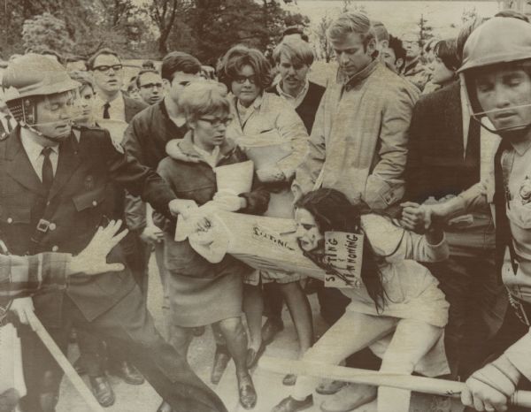 Caption on reverse of wire photo: "She Digs In--An unidentified anti-war protester with designs painted on her face and carrying sign, "Sifting and Winnowing," referring to University of Wisconsin's traditional stand in defense of academic freedom, fights removal during demonstration on campus Wednesday. Onlookers watch the action. The police won."<p>This image was published in <i>Rads, A True Story of the End of the Sixties</i>, by Tom Bates, in photo section between page 264 and 265. Caption reads, "Dow Day, October 18, 1967: Madison police detain a member of the San Francisco Mime Troupe. (<i>Courtesy</i> The Capital Times/<i>University of Wisconsin-Madison Division of Archives).</i>"

The protester is identified as Vicki Gabriner. She was not a member of the SF Mime Troupe, although the group was in town and some protesters decided to dress up.