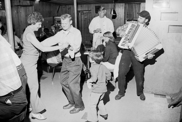 A couple dancing to accordion music at a book signing party at the Club Tavern. A young boy is dancing near them in the center, and the accordion player is standing and playing on the right. Other patrons are sitting and standing in the background. People are sitting at the bar on the left.