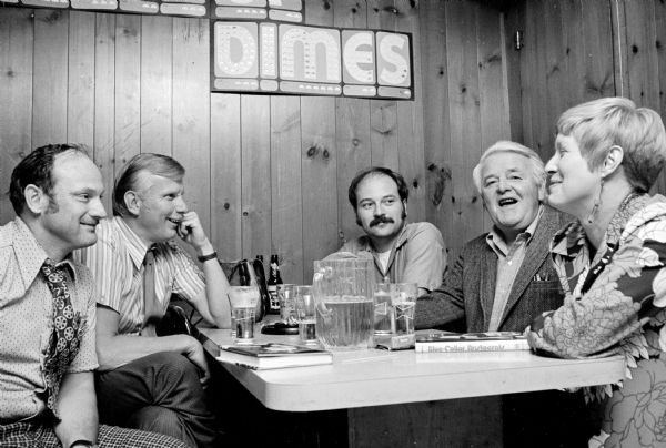 A group of three men and one woman sitting in a booth at the Club Tavern with author E.E. LeMasters, who is wearing a sport coat. Copies of the author's book, <i>Blue-Collar Aristocrats,</i> are sitting on the table during the signing party, along with beer glasses and pitchers. Overhead on the wall, which is paneled in "knotty pine," dimes are inserted in a fundraiser sign.