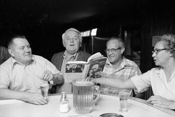 Two men and a woman discussing E.E. LeMasters' book, <i>Blue-Collar Aristocrats,</i> with the author, who is wearing a sport coat, at a book signing party at the Club Tavern. They are sitting at the bar with beer glasses and a pitcher. The woman on the right is holding the book open in front of the author.