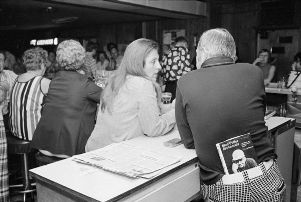 View from behind of a female patron and a man, perhaps a bartender, chatting at a book signing party at the Club Tavern. The bartender has the book, <i>Blue-Collar Aristocrats,</I> by E.E. LeMasters, tucked into the back of his plaid pants. Other patrons are sitting at the bar in the background.