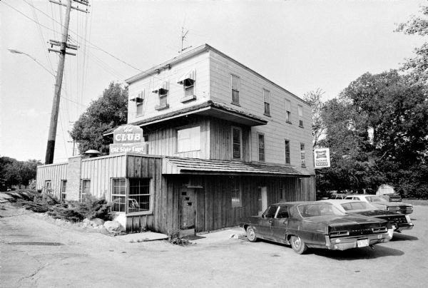 Exterior of the Club Tavern, with automobiles parked along the side. Trees, shrubs and a parking lot surround the building.