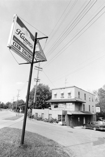 Exterior view of the Club Tavern, with a Hamm's Beer and Club Tavern electric sign with an arrow pointing to the tavern in the foreground at the entrance to the parking lot. Automobiles are parked along the right side of the building near the door. Trees, shrubs and a parking lot surround the building.
