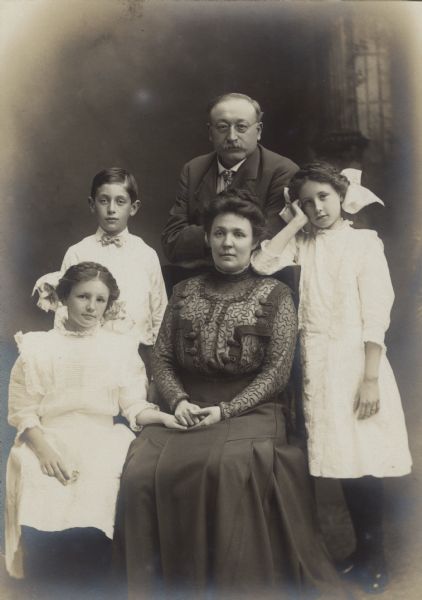 Formal studio portrait of the Berger family and a young boy in front of a painted backdrop. Names, left to right: Doris Berger, Jack Anderson, Victor Berger, Meta Berger, and Elsa Berger. Doris and Meta are sitting, holding hands. Jack, Victor and Elsa are standing, with Victor resting his arms on the back of Meta's chair.